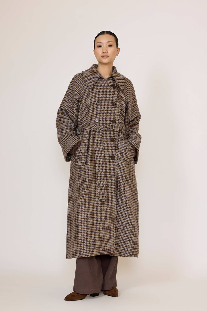Person wearing a long brown checkered coat and brown pants underneath on a white background