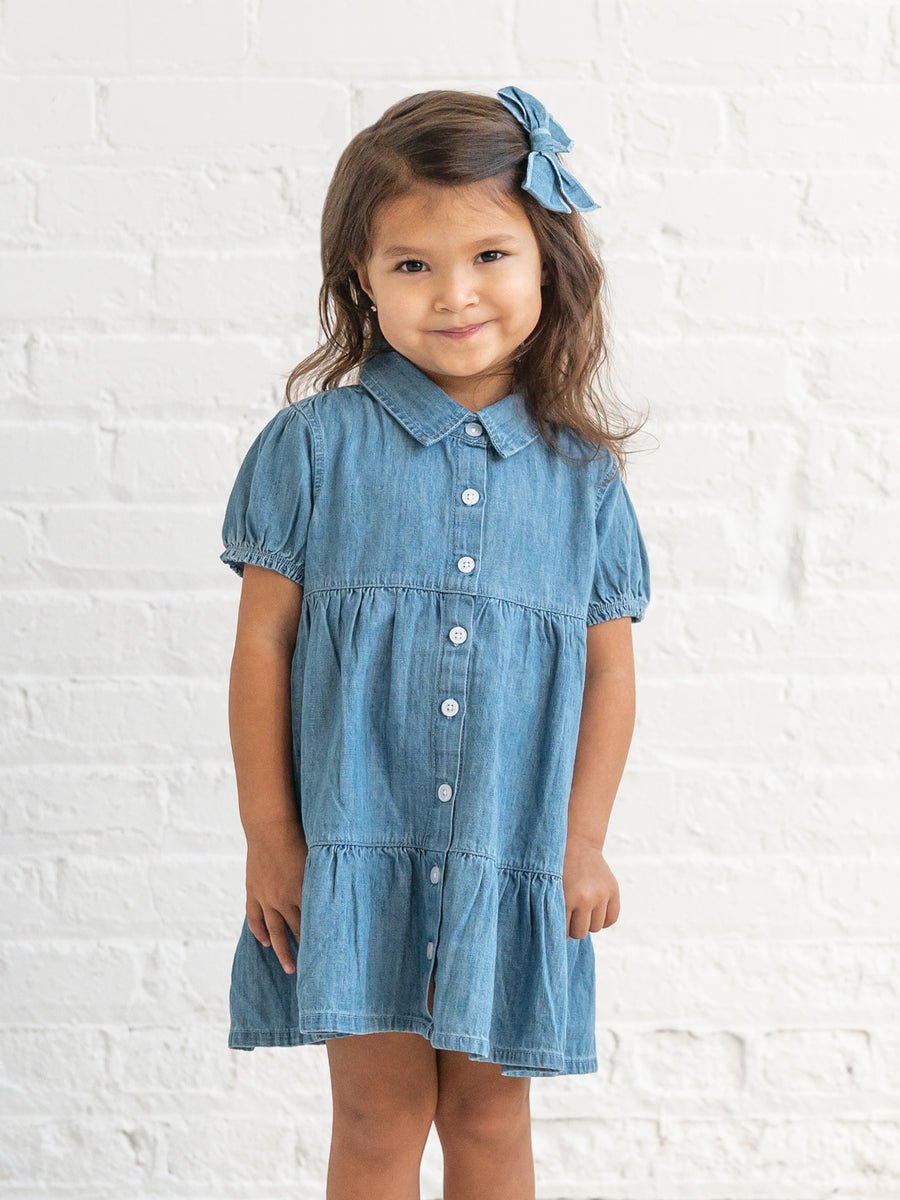 Young girl wearing a chambray dress with a white brick wall background