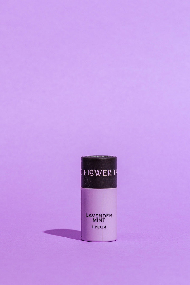 A single tube of Good Flower Farm Lavender Mint Lip Balm stands upright on a solid lavender-purple background. The lip balm tube is a pale lavender color with a black cap, and the brand name &