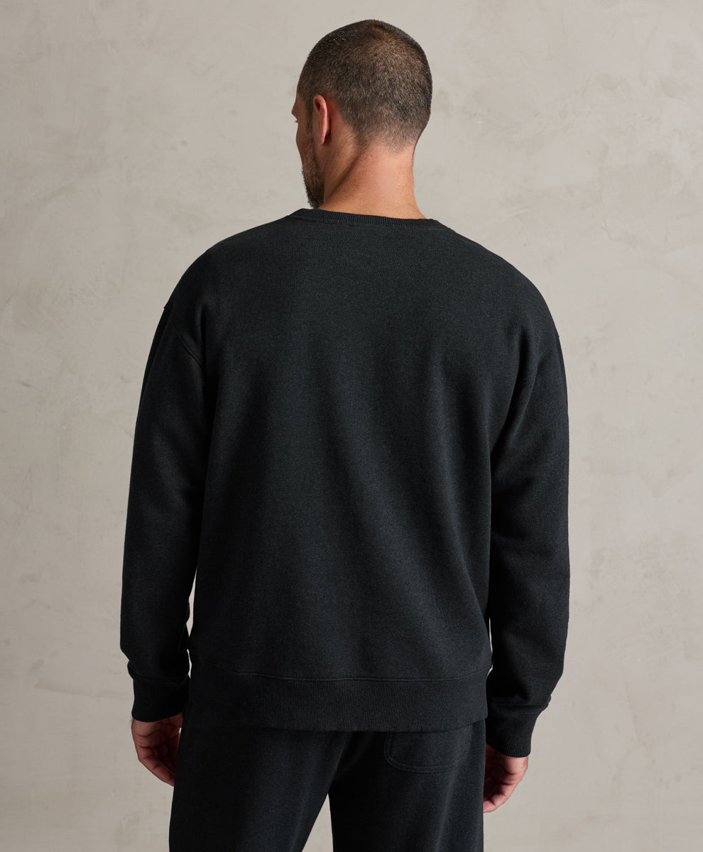 A man viewed from behind wearing a charcoal heather crewneck sweatshirt and matching sweatpants against a textured light gray wall.