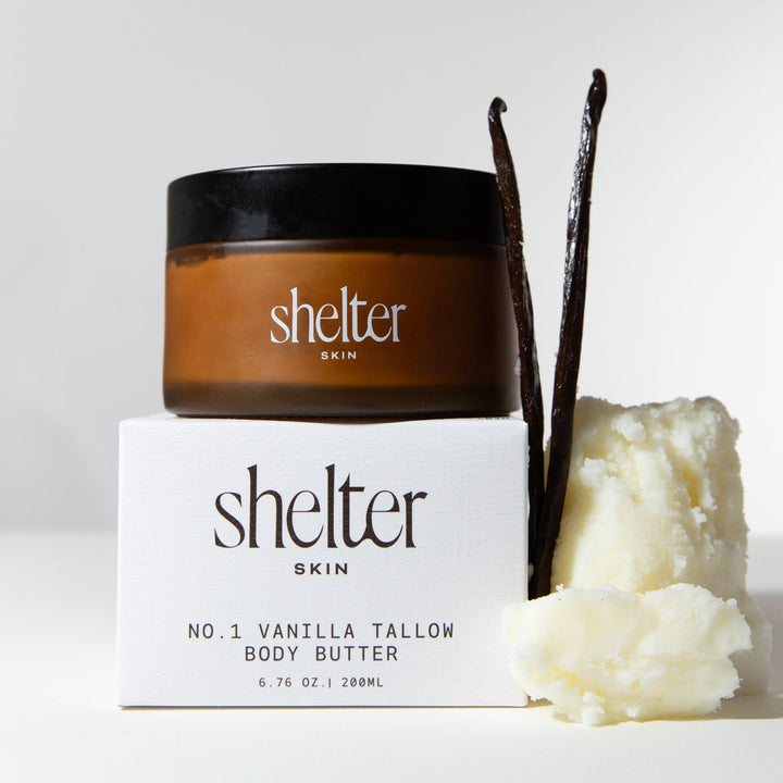Shelter Skin No. 1 Vanilla Tallow Body Butter in an amber jar with a black lid, displayed on top of its white box with vanilla bean pods and vanilla butter beside it.