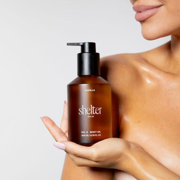 A woman holding a bottle of Shelter Body Oil in Vanilla scent. The bottle is amber brown with a black pump dispenser and white text.