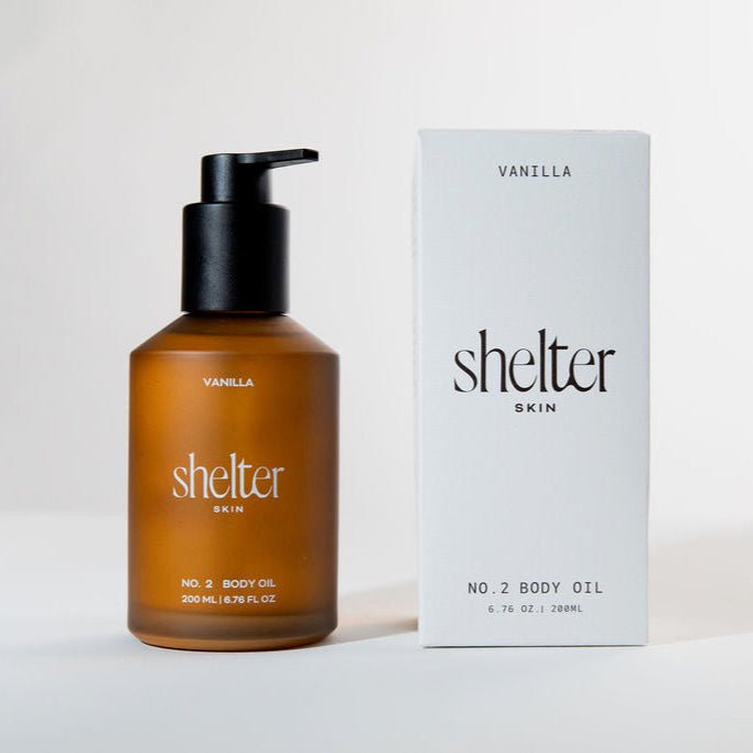 Shelter Skin No. 2 Body Oil in Vanilla scent, shown in an amber glass bottle with a black pump next to its white packaging box.