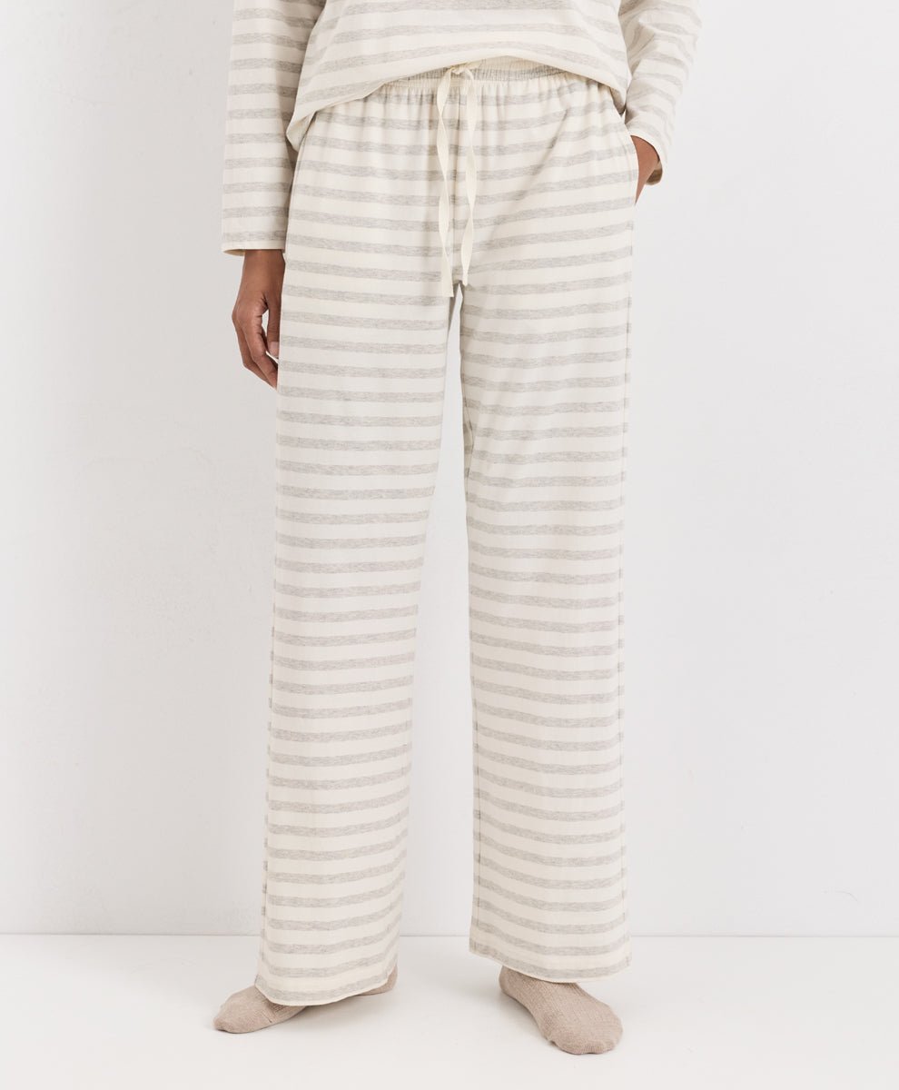 A person wearing loose, wide-leg pajama pants with thin horizontal beige and off-white stripes, paired with a matching long-sleeve top and socks, photographed against a plain light background.