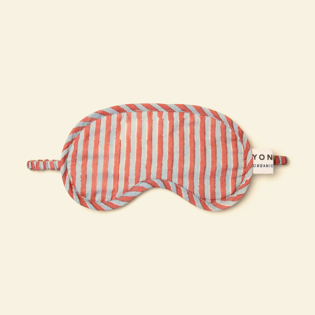 A striped sleep mask with vertical coral pink and light blue stripes, featuring a small white tag on the side that reads &