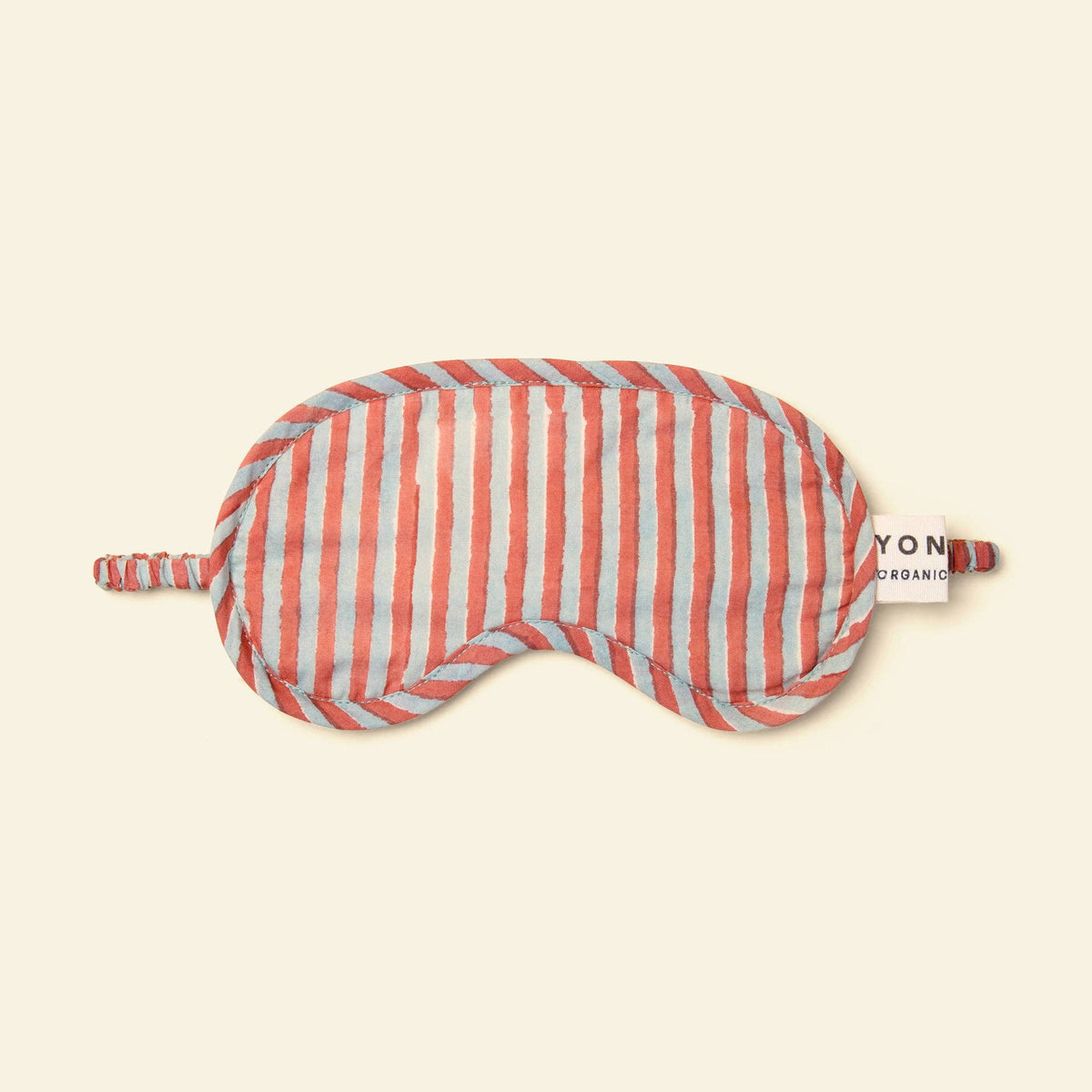 A striped sleep mask with vertical coral pink and light blue stripes, featuring a small white tag on the side that reads 'Yonder ORganic'. The mask is laid flat on a solid cream-colored background.