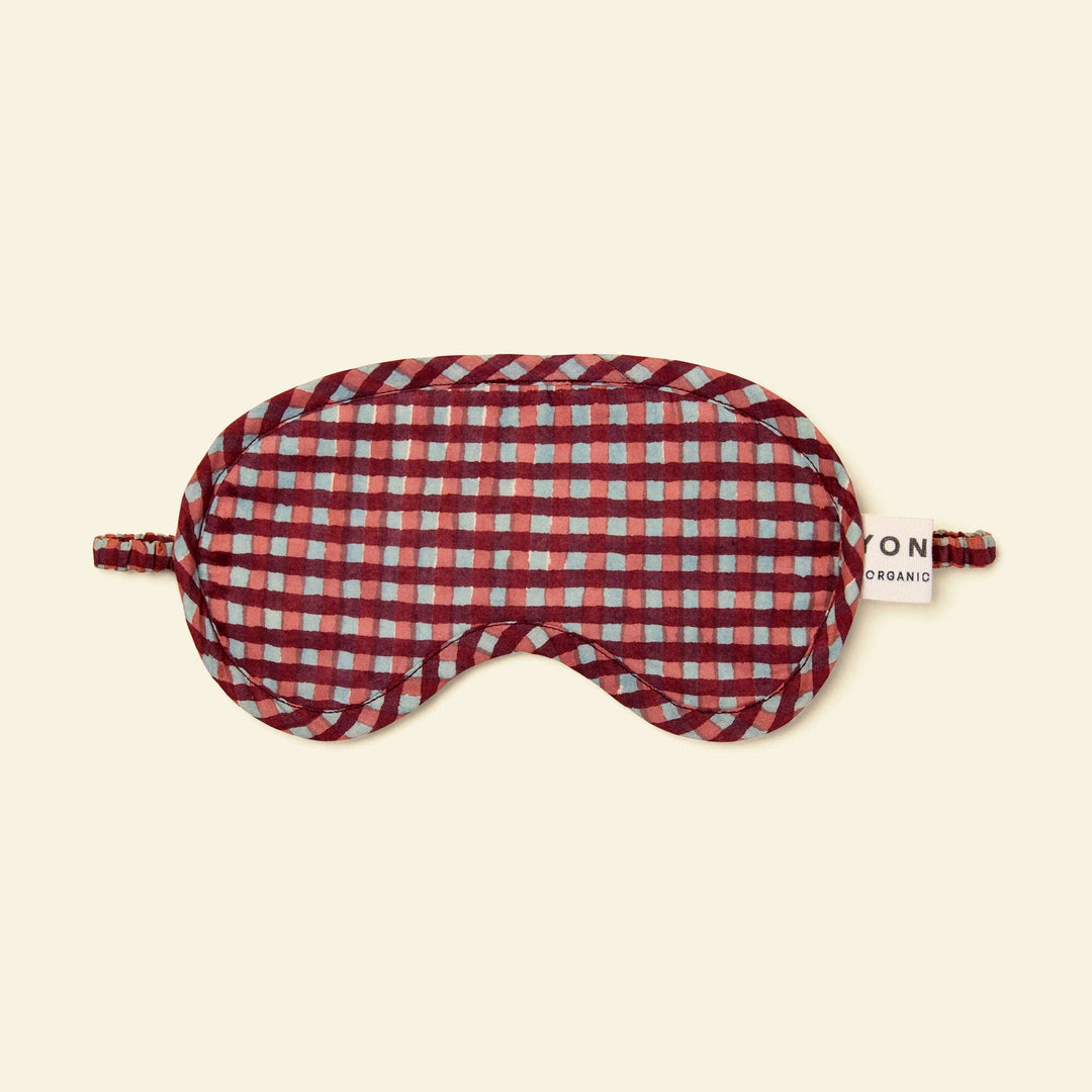 A red and white gingham patterned sleep mask with adjustable ear loops and a small white brand tag reading &