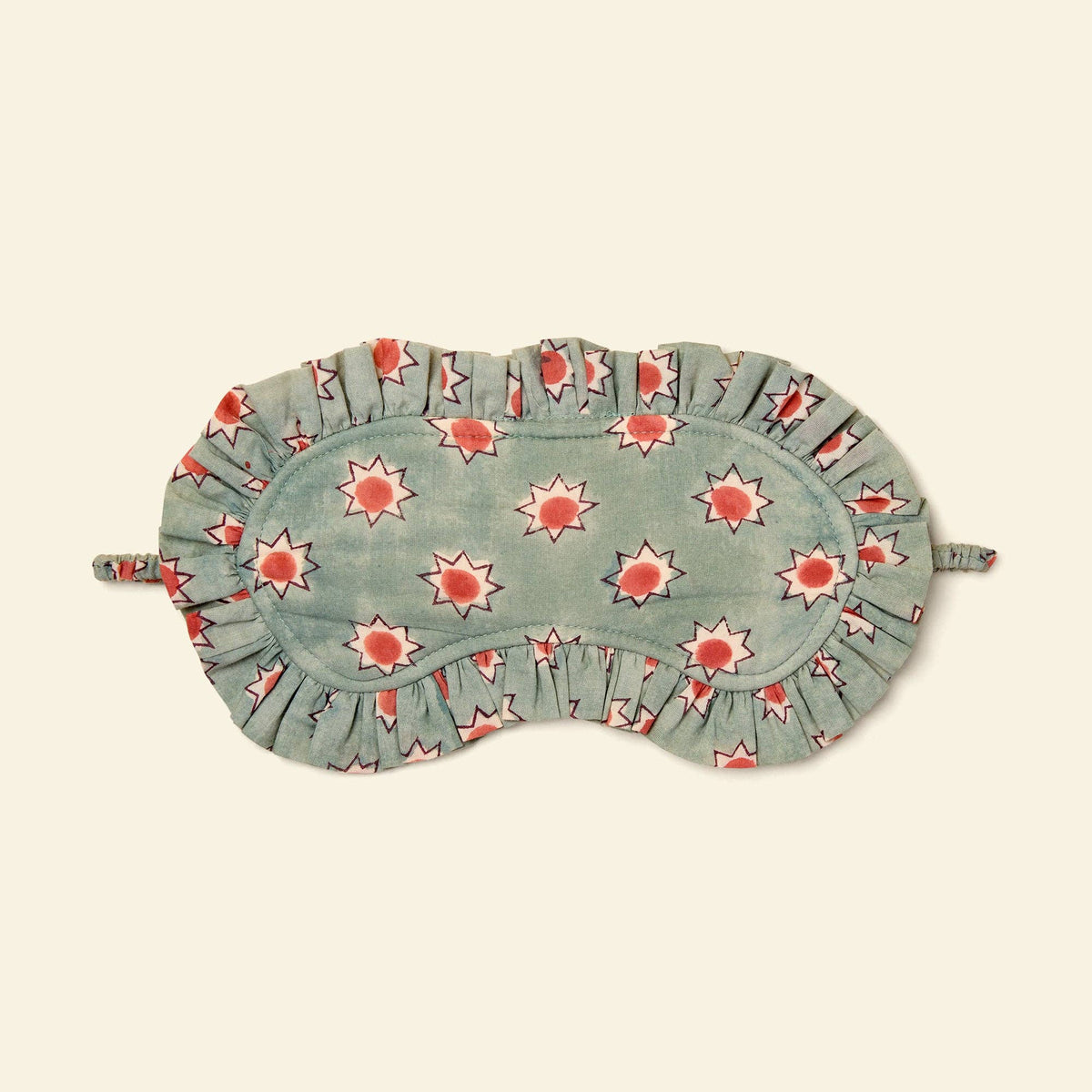 A fabric sleep mask with a ruffled edge, featuring a sage green background and a repeating pattern of red sunflowers with white outlines, shown on a solid cream background.