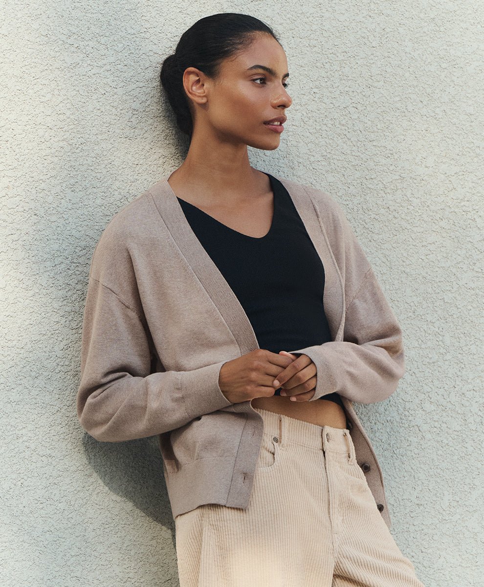 A woman wearing a soft umber heather cardigan over a black V-neck top and beige corduroy trousers, leaning against a textured light gray wall.