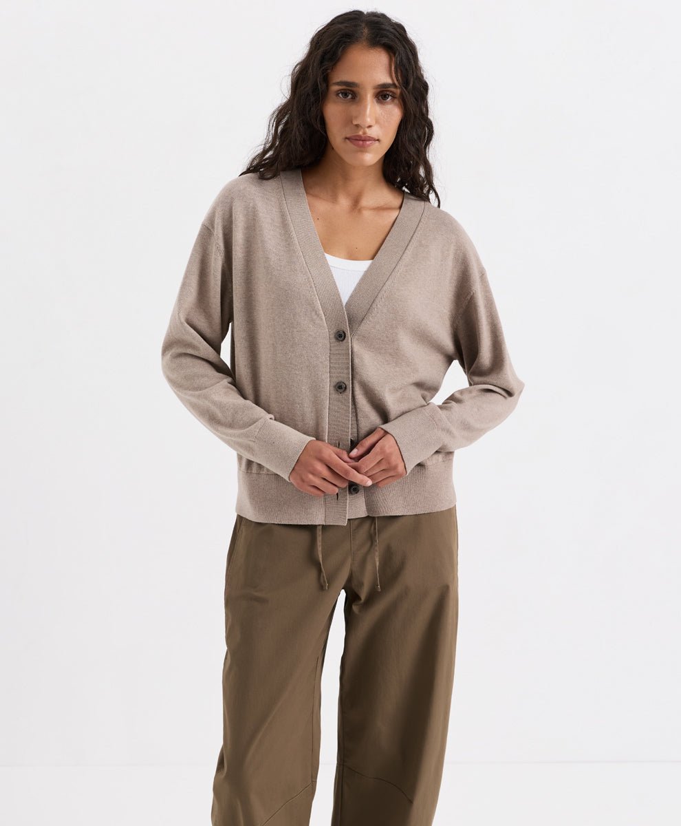 A woman modeling Pact's soft umber heather knit cardigan with dark buttons over a white top and brown drawstring trousers, shot against a clean white studio background.