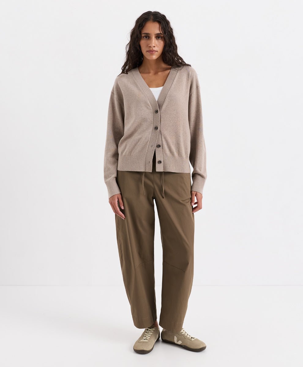A woman stands against a plain white studio background wearing a soft umber heather knit cardigan over a white top, loose brown pants, and taupe sneakers.