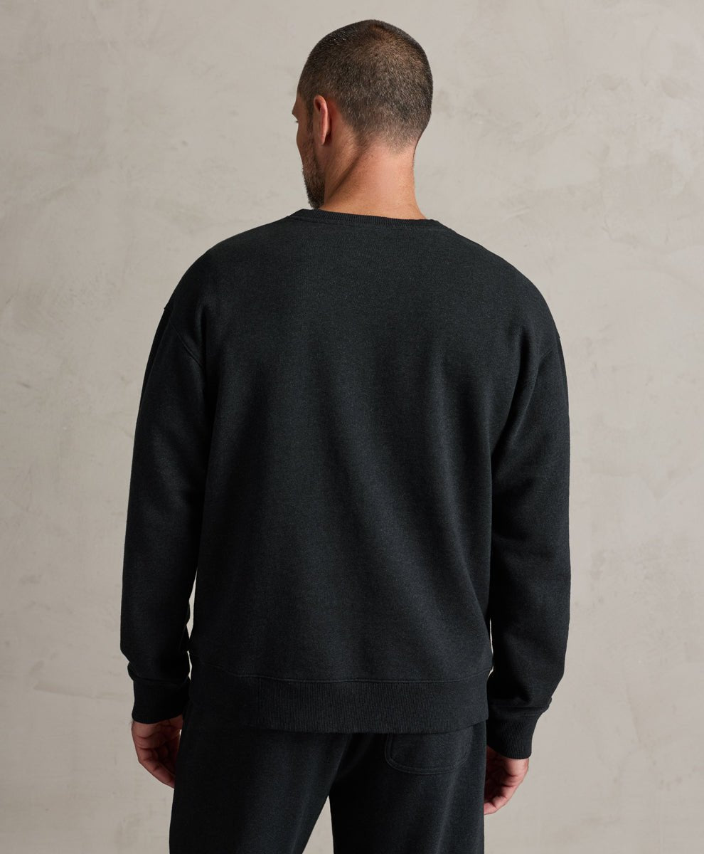 A man viewed from behind wearing a charcoal heather crewneck sweatshirt and matching sweatpants against a textured light gray wall.