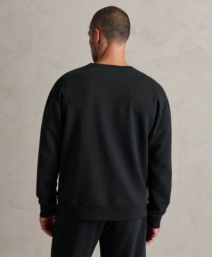 A man viewed from behind wearing a charcoal heather crewneck sweatshirt and matching sweatpants against a textured light gray wall.