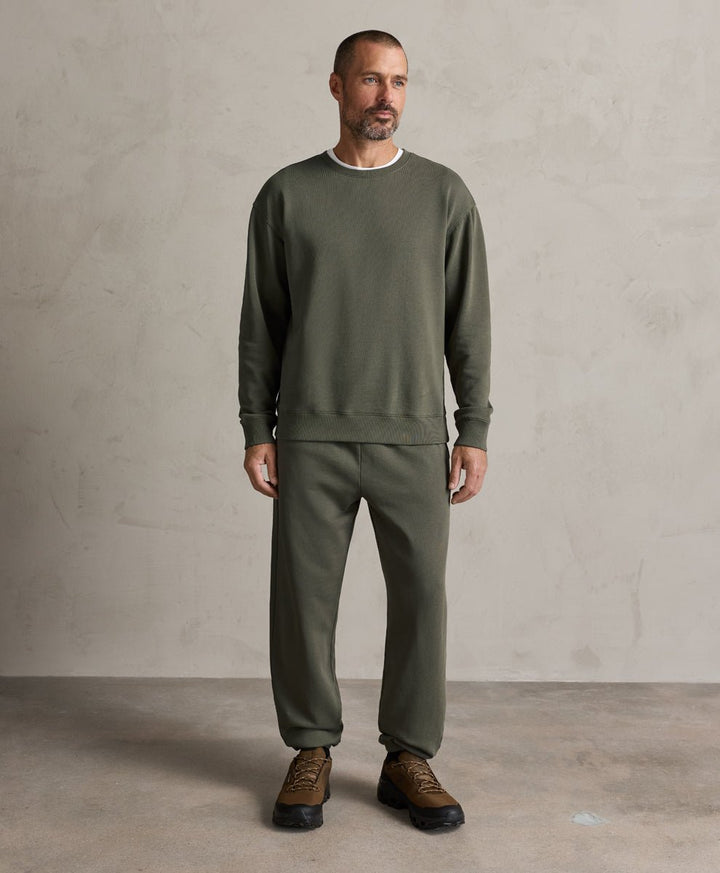 A man wearing a dusted green crewneck sweatshirt and matching sweatpants, paired with brown leather sneakers, standing against a textured light gray studio wall.