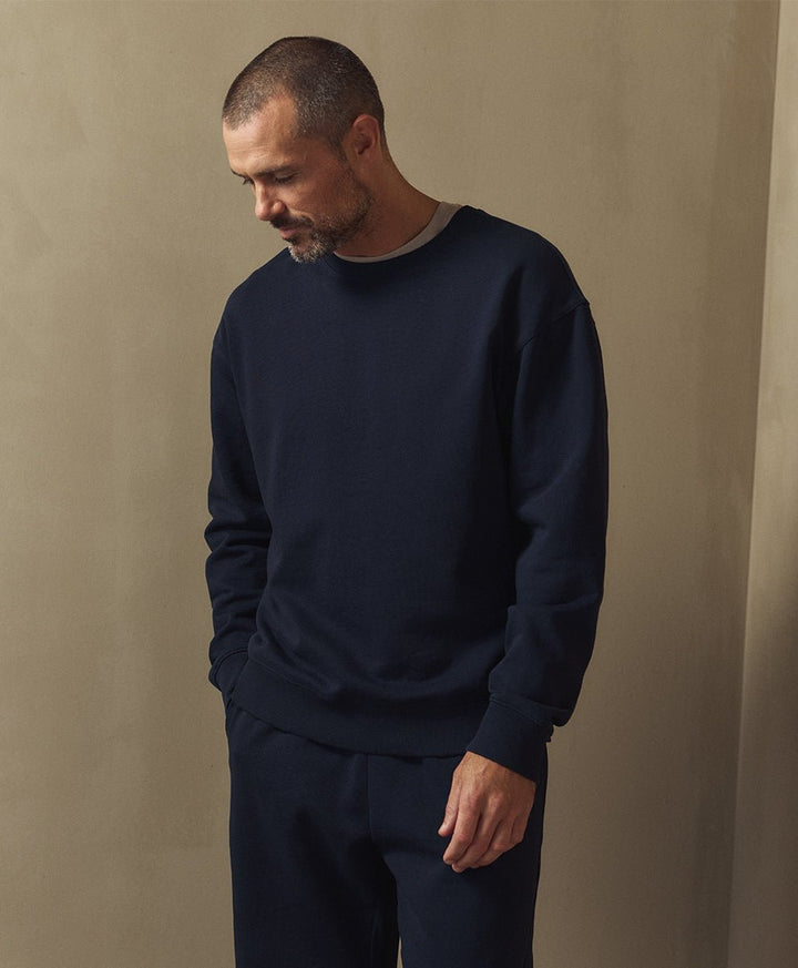 A man wearing a dark navy crewneck sweatshirt and matching navy pants, standing against a textured beige wall.