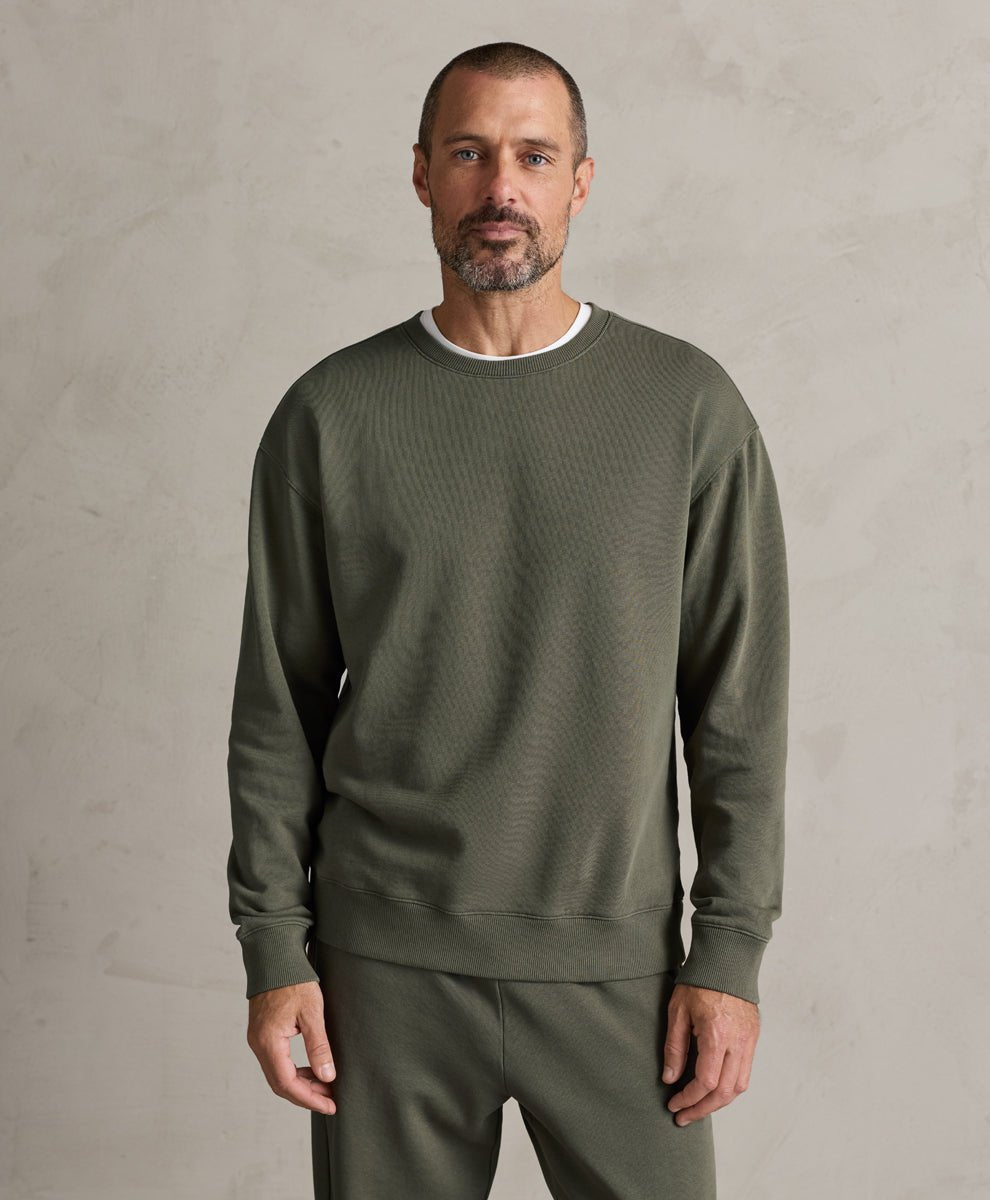 A man wearing a Pact dusted green ribbed crewneck sweatshirt and matching sweatpants, standing against a textured light gray wall.