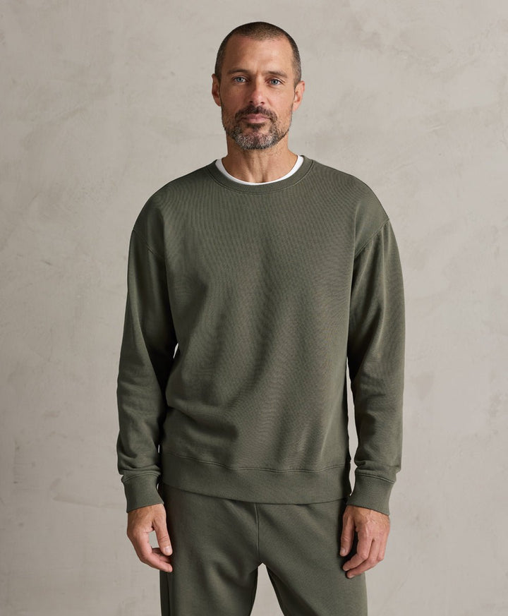 A man wearing a Pact dusted green ribbed crewneck sweatshirt and matching sweatpants, standing against a textured light gray wall.