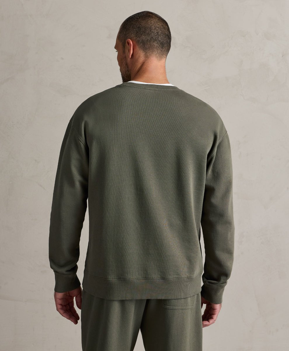 A man viewed from behind wearing an dusted green crewneck sweatshirt and matching sweatpants against a textured light gray wall.