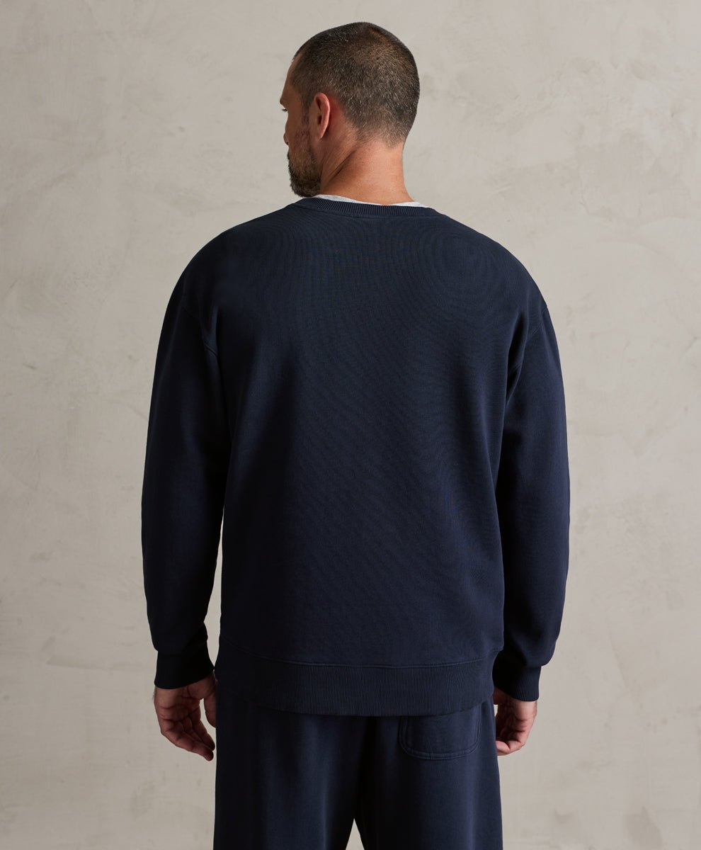 A man viewed from behind wearing a dark navy blue ribbed knit crewneck sweatshirt and matching navy pants, standing against a textured light beige wall.
