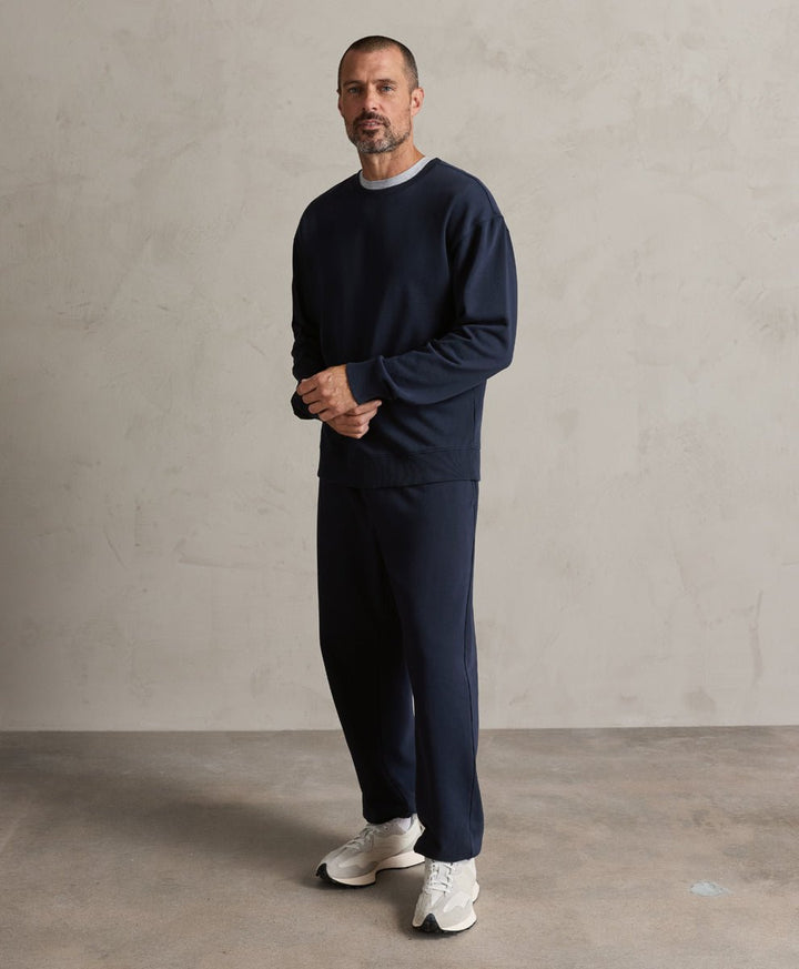 A man wearing a dark navy blue knit crewneck sweatshirt and matching navy pants, paired with white sneakers, standing against a textured light gray wall.