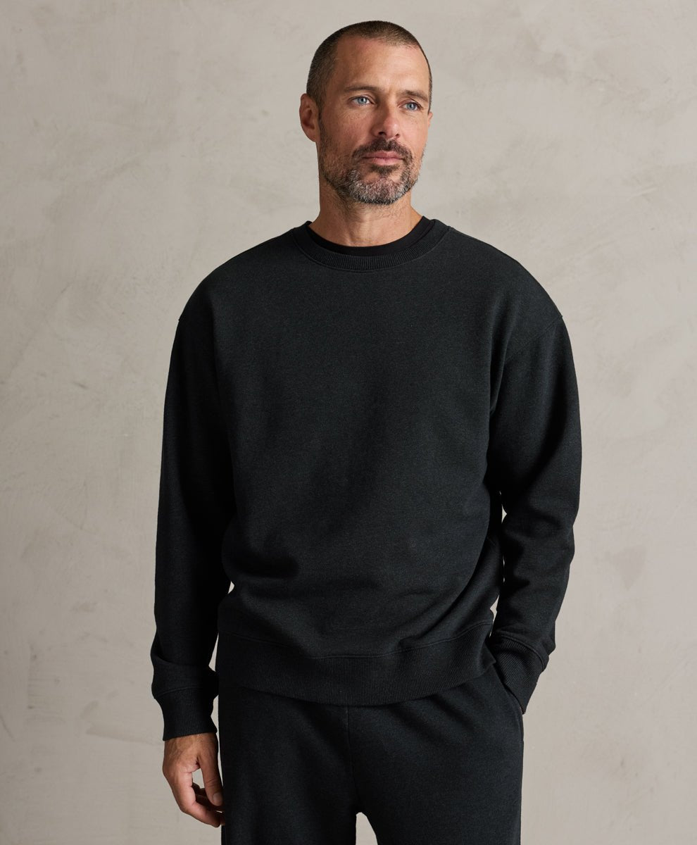 A man wearing a Pact charcoal heather long-sleeve crewneck sweater and matching black pants, standing against a textured light gray wall.