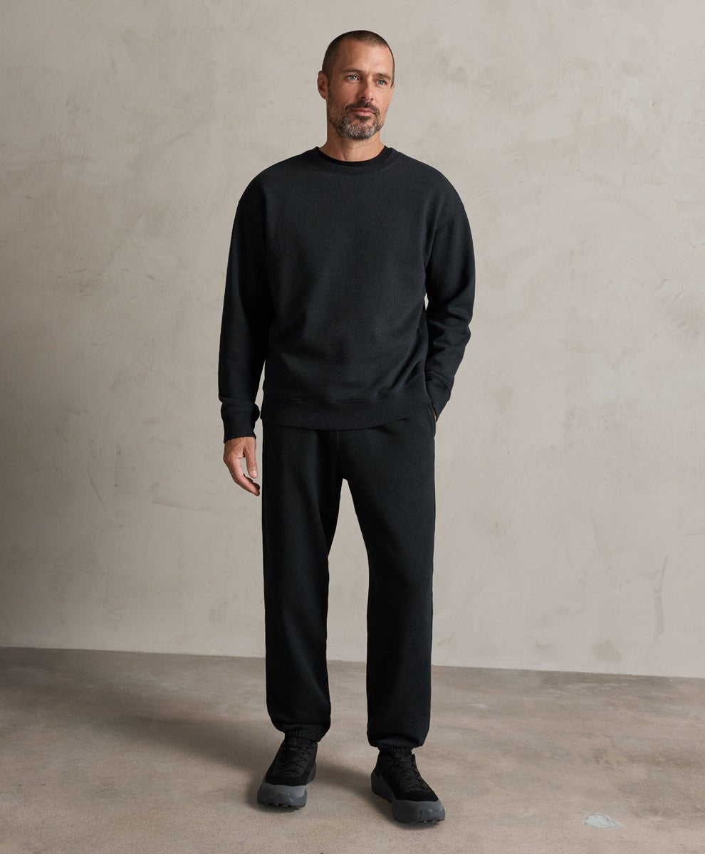 A man wearing a pact charcoal heather crewneck sweatshirt and matching black sweatpants, standing against a textured light gray studio wall.