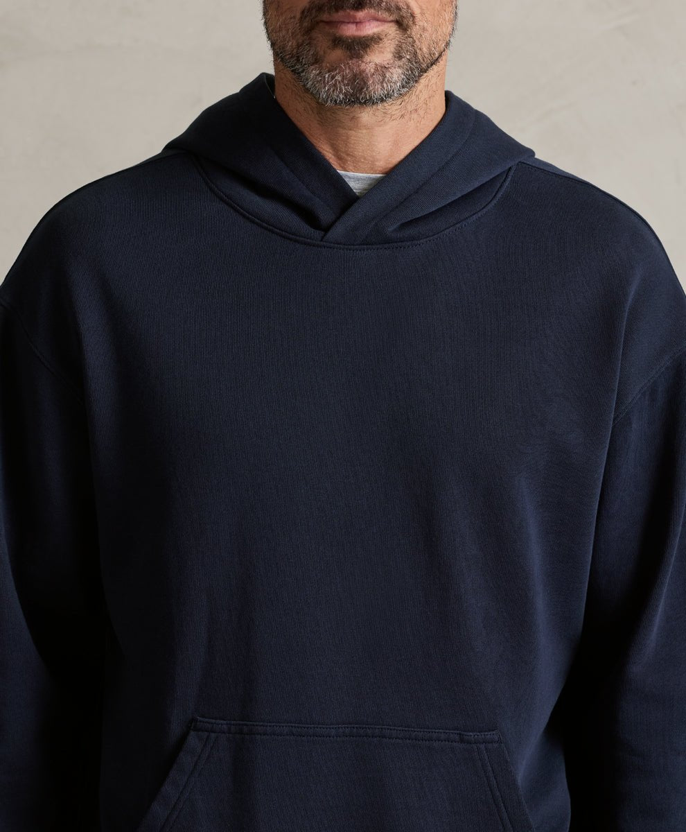 A close-up of a man wearing a dark navy blue pullover hoodie with a kangaroo pocket, shown from the chest up against a neutral textured wall.