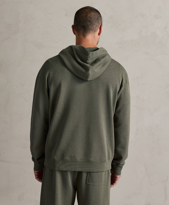 A man viewed from behind wearing a dusted green hooded sweatshirt and matching sweatpants against a textured light gray wall.