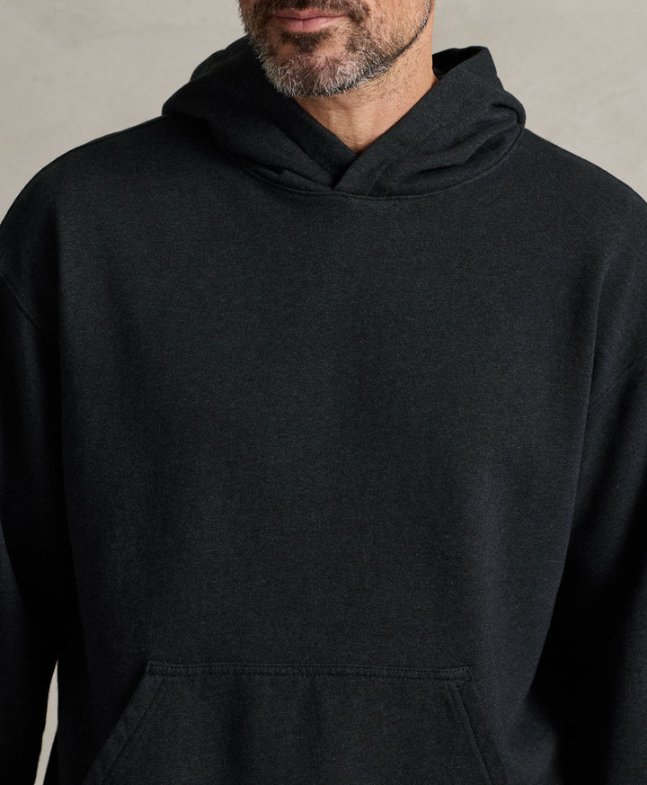 A close-up of a man wearing a charcoal heather pullover hoodie with a front kangaroo pocket, set against a neutral textured background.