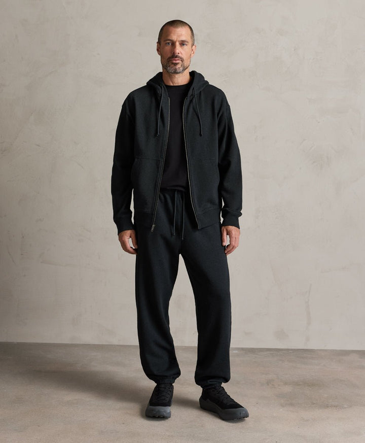 A man wearing a Pact charcoal heather zip-up hoodie, sweatpants, and black sneakers stands against a textured light gray concrete wall.