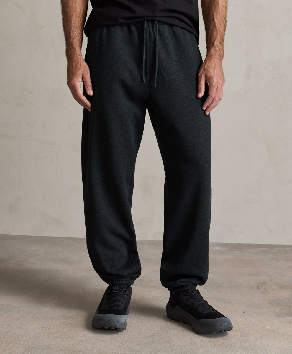A person wearing charcoal heather pact jogger-style sweatpants with a drawstring waist, shown from the chest down in a studio setting with a textured light gray wall and concrete floor.