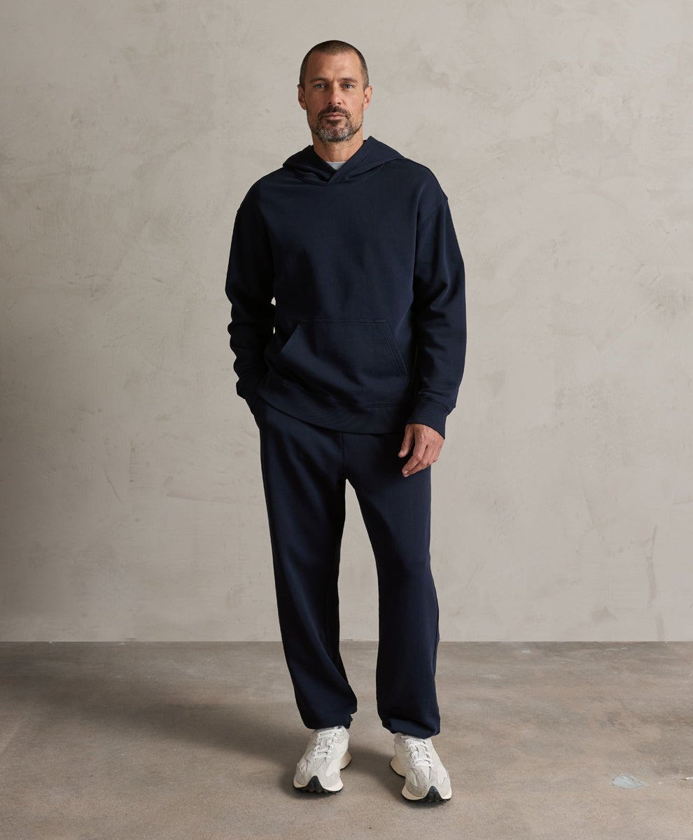 A man wearing a matching navy blue hoodie and sweatpants set with white sneakers, standing against a textured light gray wall.