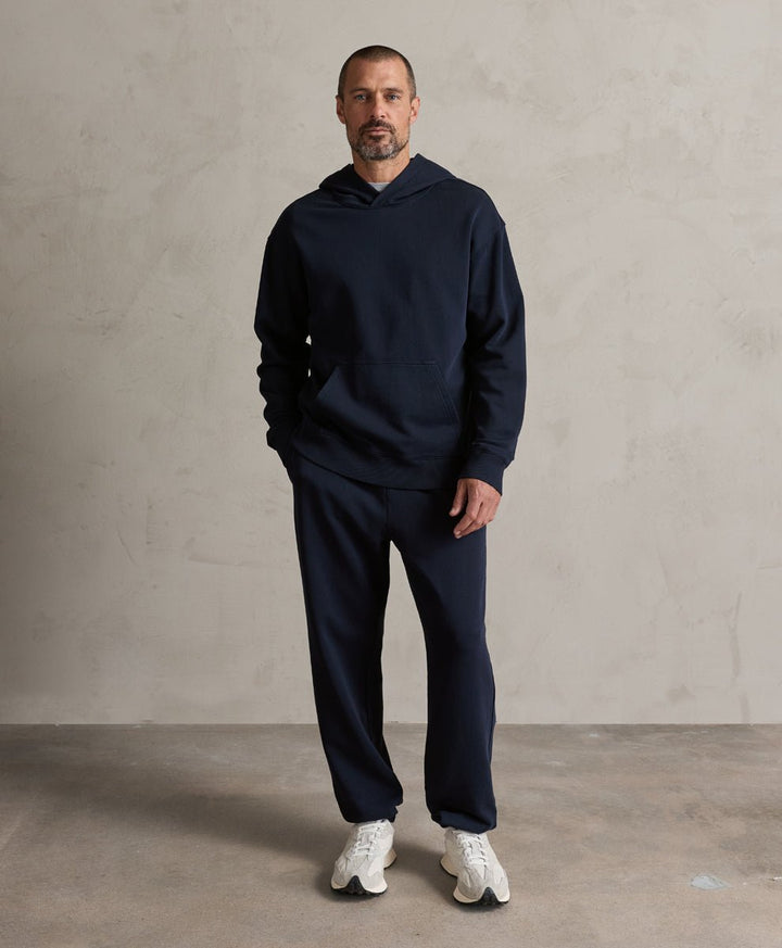A man wearing a matching navy blue hoodie and sweatpants set with white sneakers, standing against a textured light gray wall.