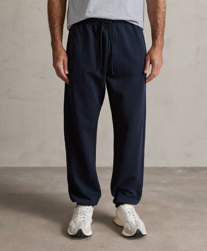A person wearing pact dark navy drawstring sweatpants, a light gray t-shirt, and white sneakers, standing against a neutral concrete-textured wall.