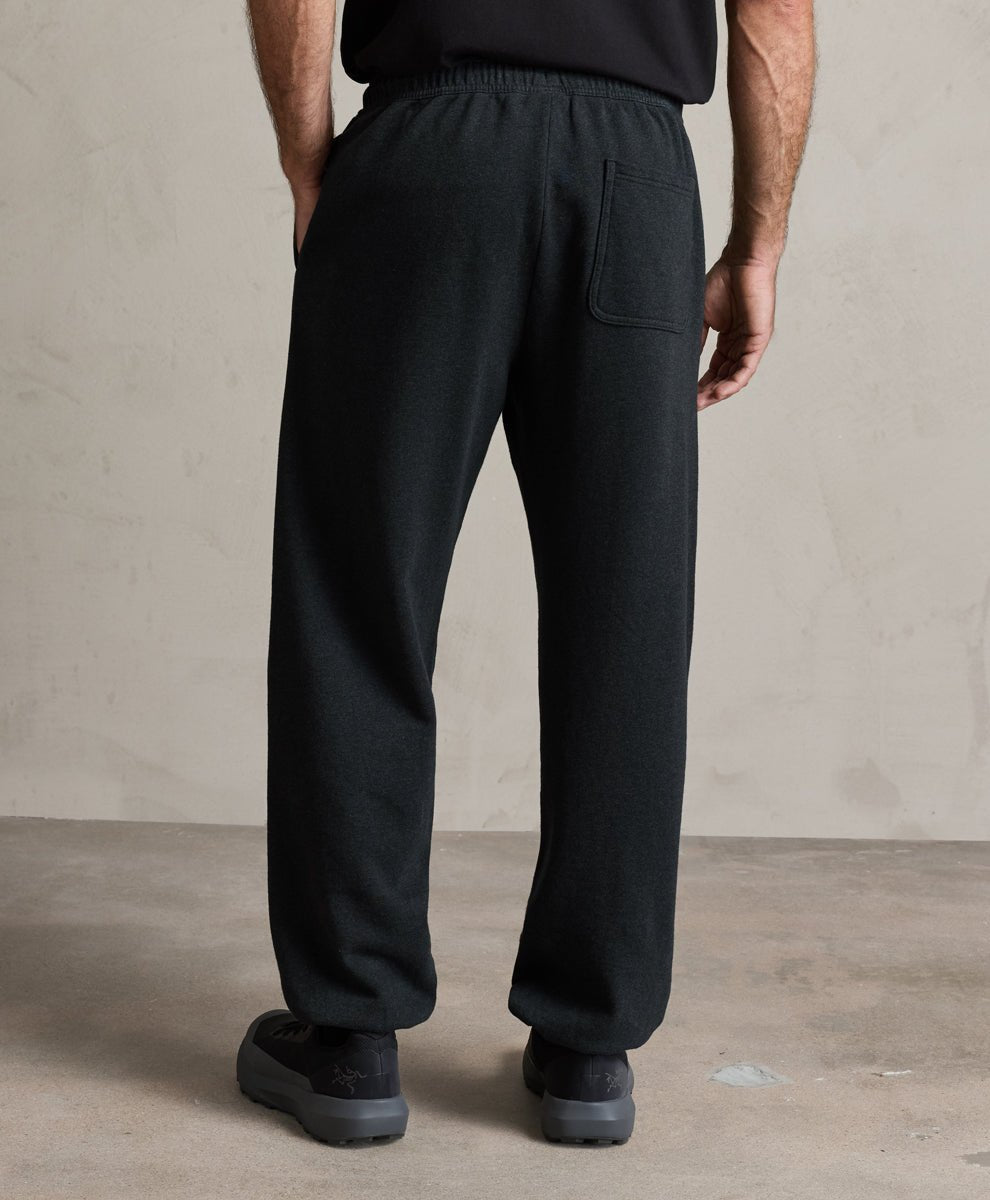 A person shown from the back wearing loose charcoal heather drawstring sweatpants with a side pocket, paired with a black top and black shoes, standing against a textured beige wall and concrete floor.