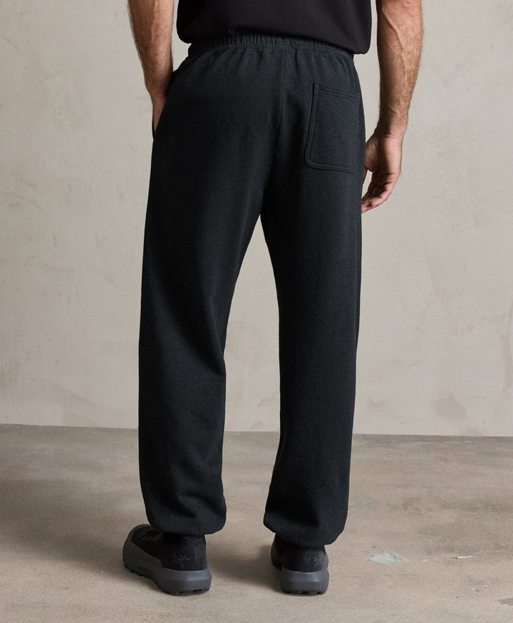 A person shown from the back wearing loose charcoal heather drawstring sweatpants with a side pocket, paired with a black top and black shoes, standing against a textured beige wall and concrete floor.