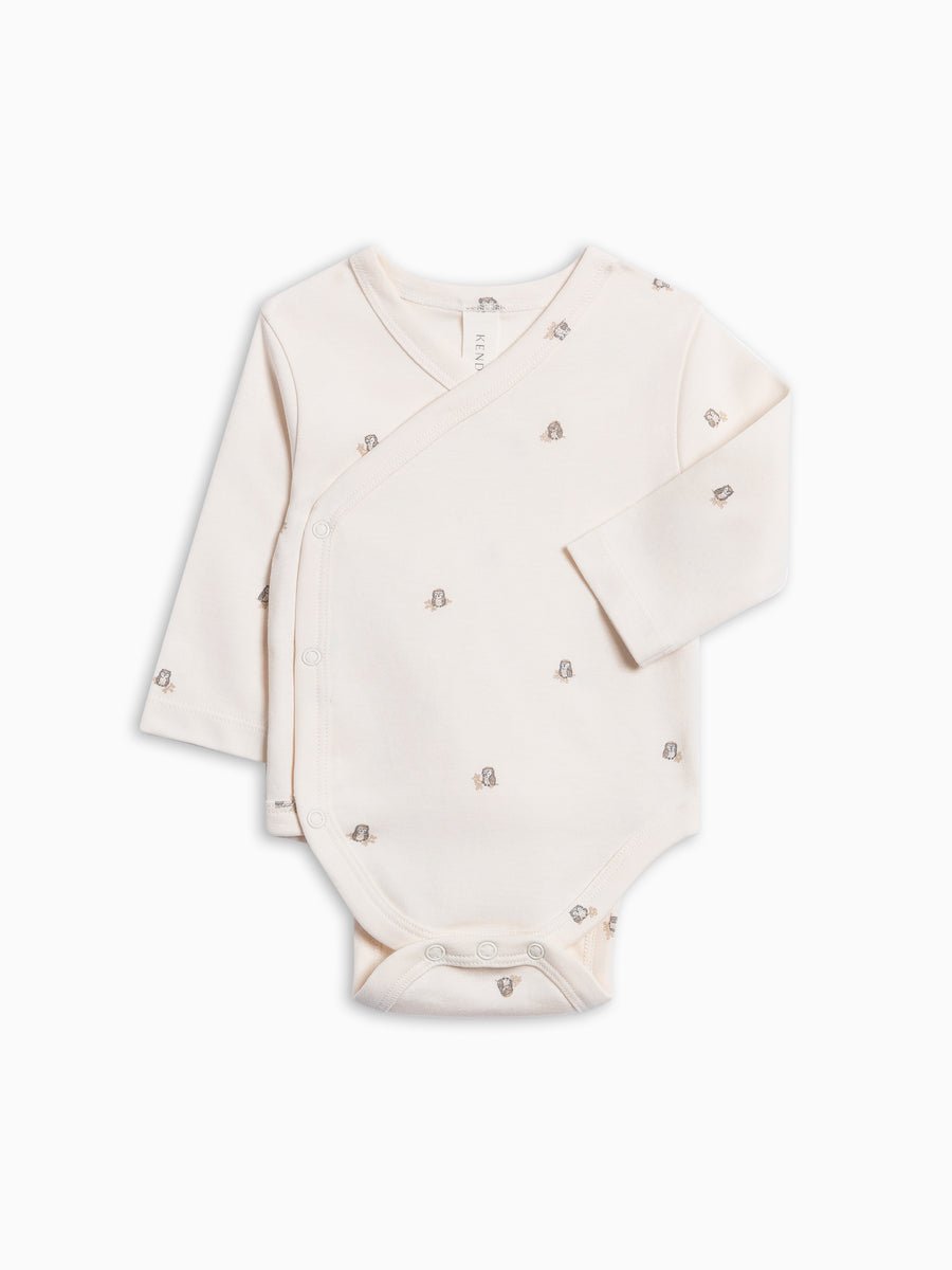 A long-sleeve baby bodysuit in a warm cream color, featuring a small repeating pattern of tiny owl illustrations and snap closures at the shoulder, laid flat on a white background.