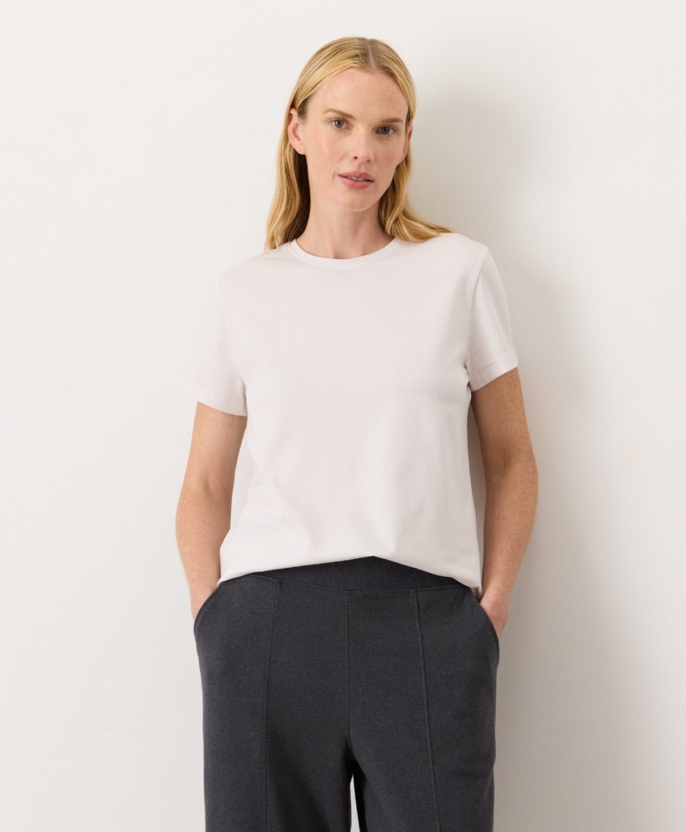 Woman wearing a Pact cream Organic Luxe Essential Crewneck Tee
 and dark pants against a plain background