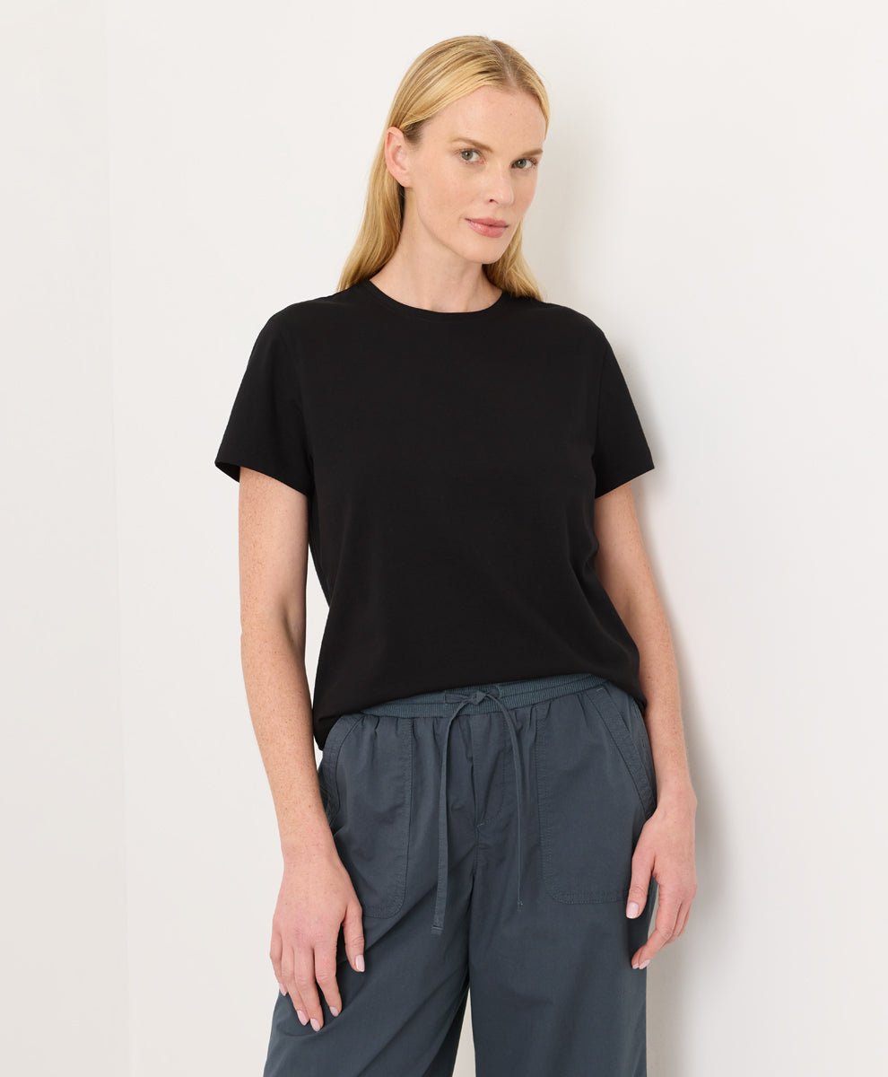 Woman wearing a Pact black Organic Luxe Essential Crewneck Tee and dark pants against a plain background
