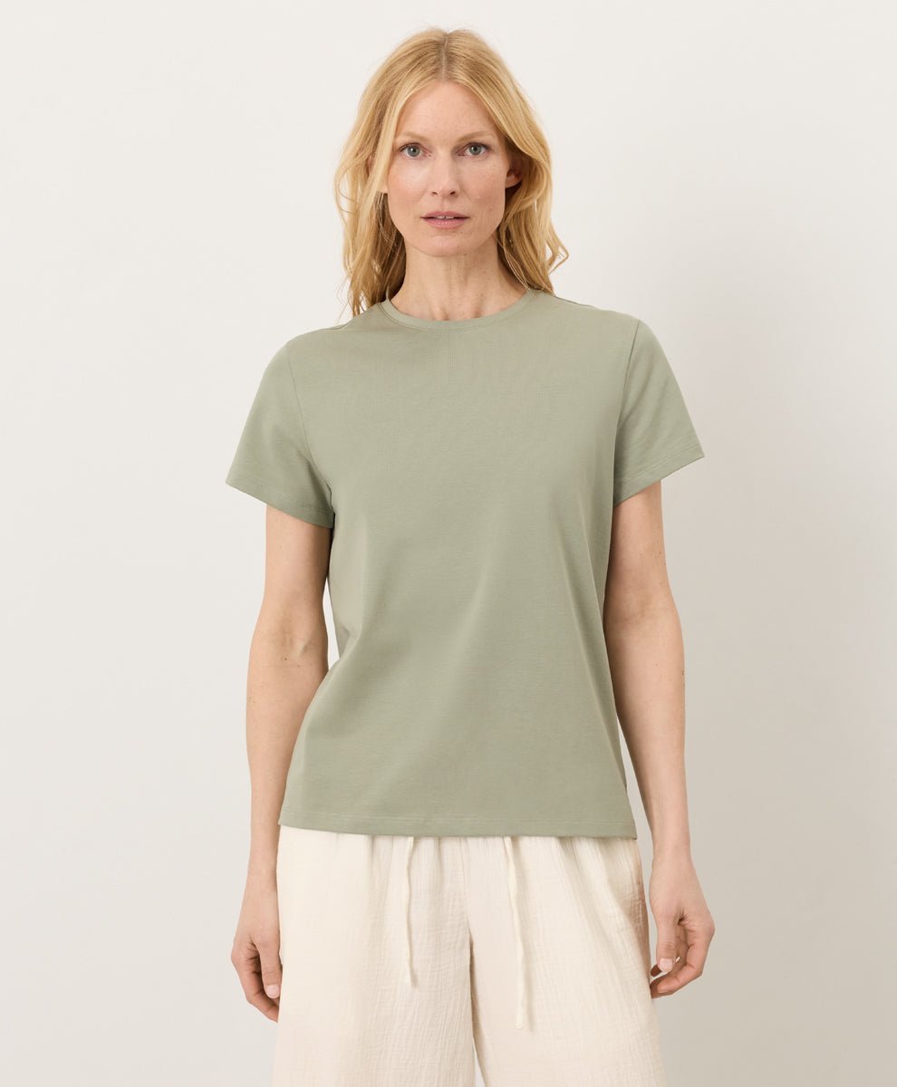 Woman wearing a Pact seagrass Organic Luxe Essential Crewneck Tee and cream pants against a plain background