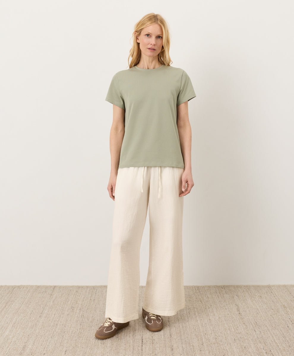 Woman wearing a Pact seagrass Organic Luxe Essential Crewneck Tee and cream pants against a plain background
