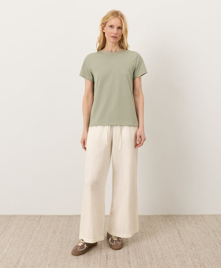 Woman wearing a Pact seagrass Organic Luxe Essential Crewneck Tee and cream pants against a plain background