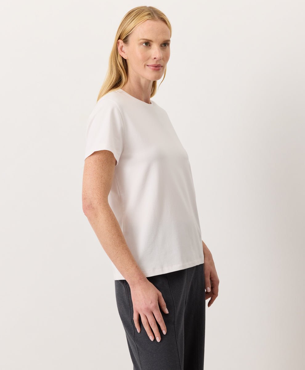 Woman wearing a Pact cream Organic Luxe Essential Crewneck Tee
 and dark pants against a plain background