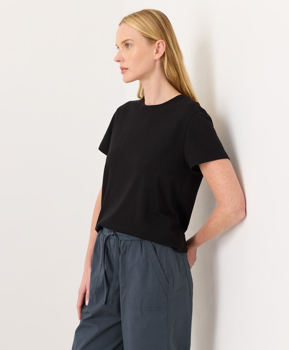 Woman wearing a Pact black Organic Luxe Essential Crewneck Tee
 and dark pants against a plain background