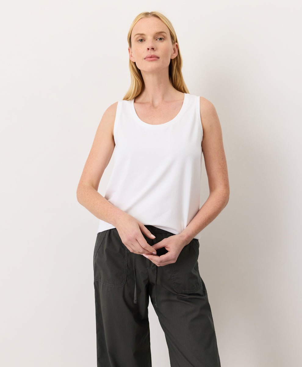 Woman wearing a white Pact Organic Luxe Essential Scoop Neck Tank and dark pants against a white background