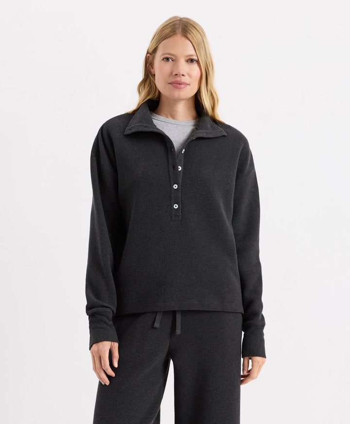 A woman modeling a Pact dark charcoal heather long-sleeve quarter-placket henley top with white buttons, paired with matching dark sweatpants and a light gray undershirt, against a plain white studio background.