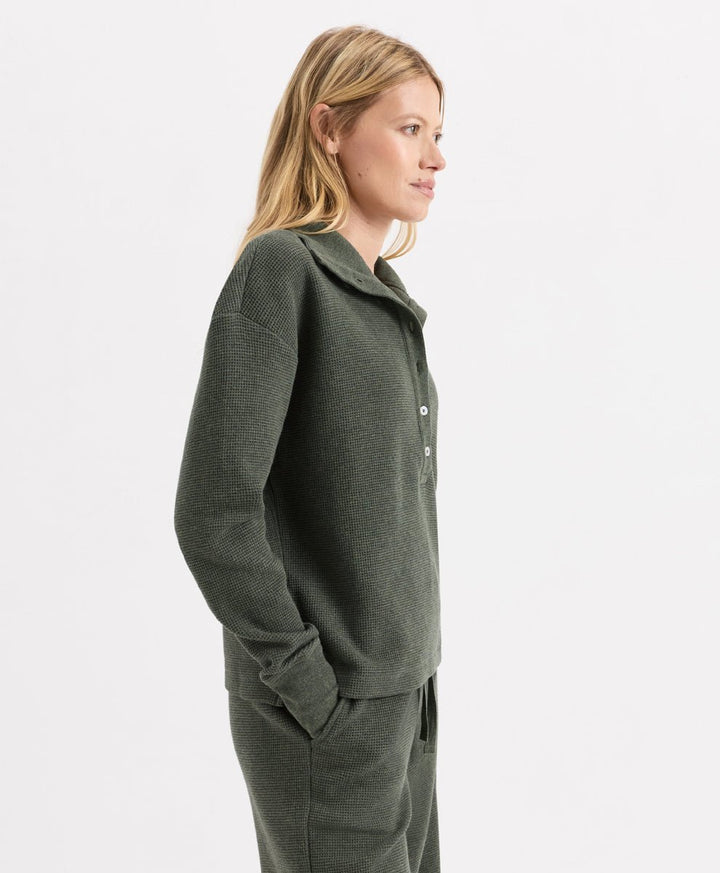 A woman in profile wearing a dusted green heather knit matching set, featuring a long-sleeve henley and coordinating pants, against a plain light gray studio background.