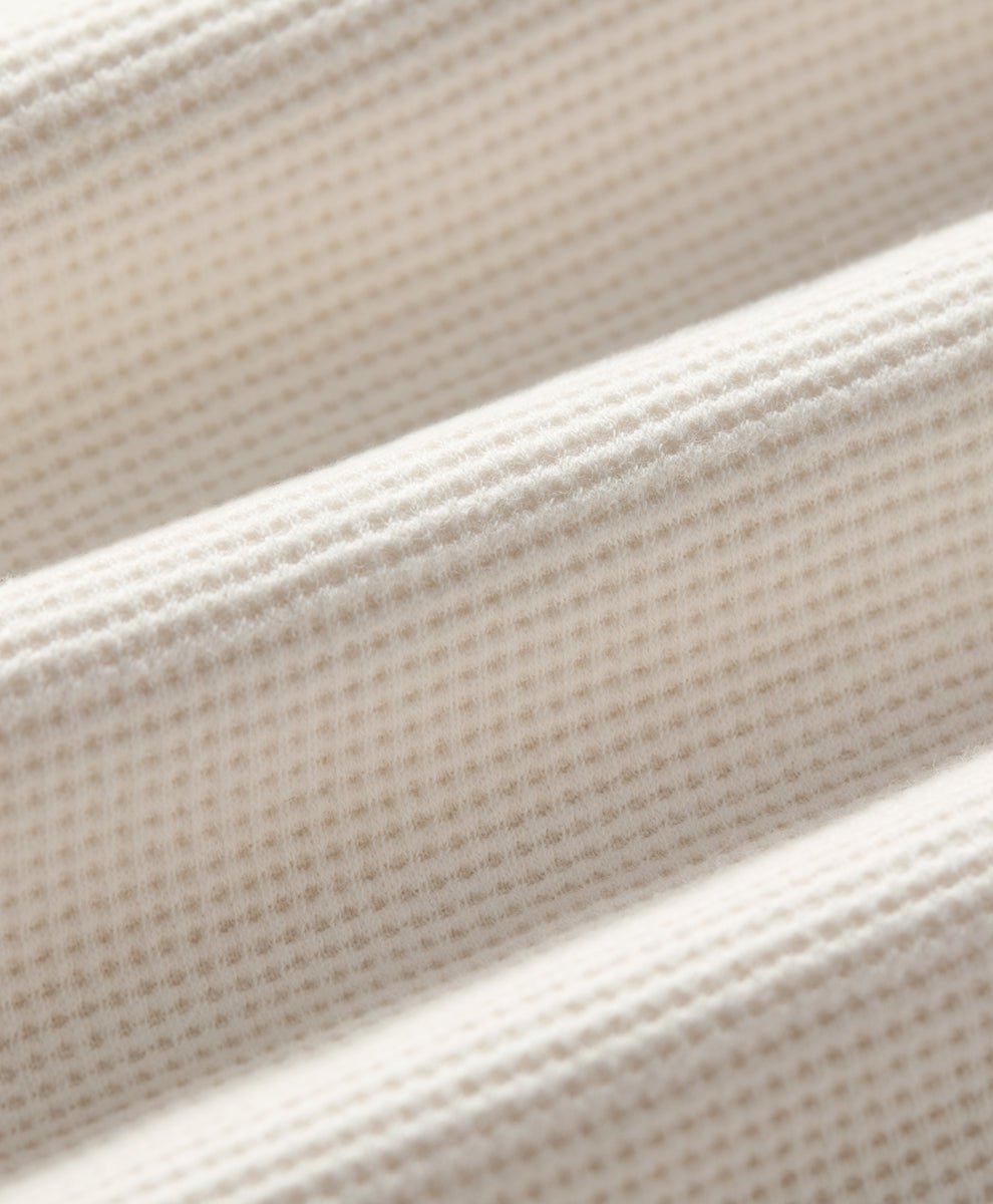 Close-up of a soft ivory-colored textured fabric with a fine waffle or grid-like weave, shown in soft diagonal folds.