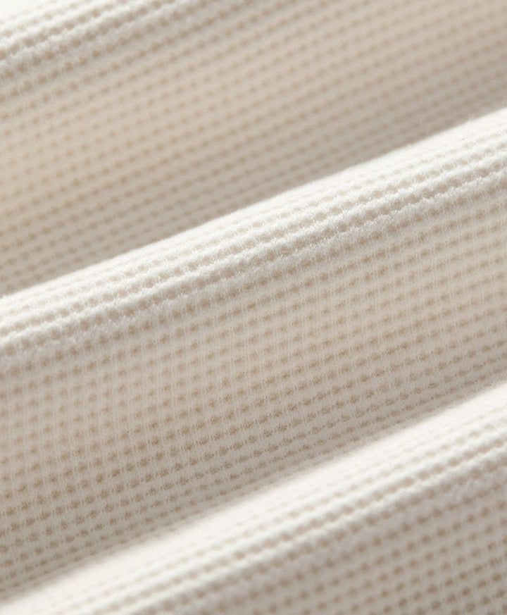 Close-up of a soft ivory-colored textured fabric with a fine waffle or grid-like weave, shown in soft diagonal folds.