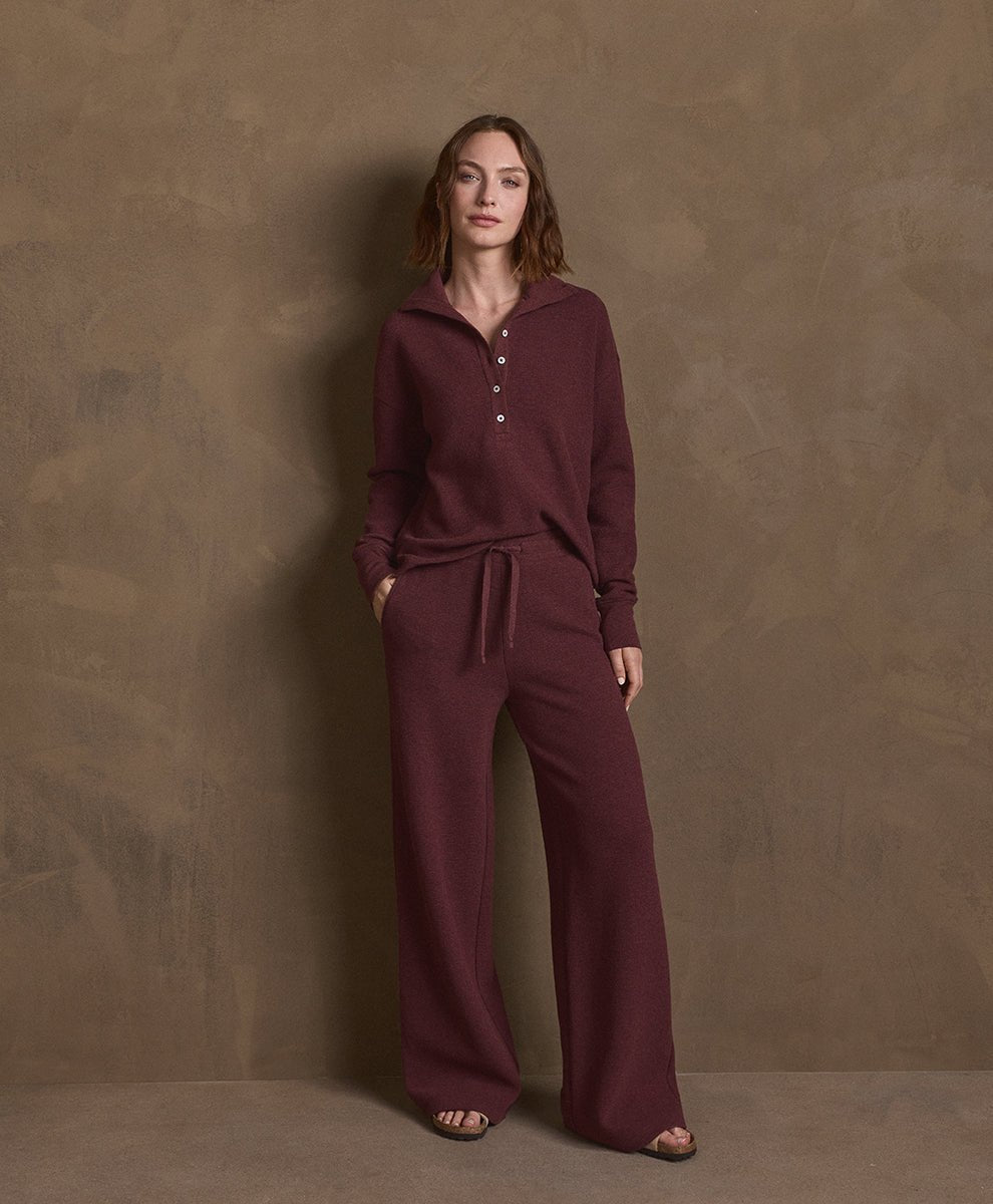 A woman stands against a textured brown studio backdrop wearing a matching merlot heather knit loungewear set consisting of a long-sleeve henley top with a drawstring and wide-leg pants.